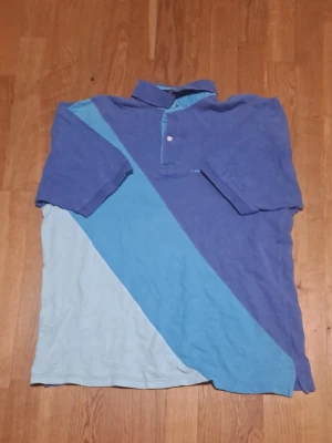 Blå och ljusblå polotopp i bomull - Säljer pikétröja i blå och ljusblå färgblock från Tommy Hilfiger i 100% bomull. Tröjan har klassisk krage, korta ärmar och knäppning med två knappar vid halsen. Den övre knappen är sönder men går att stänga.