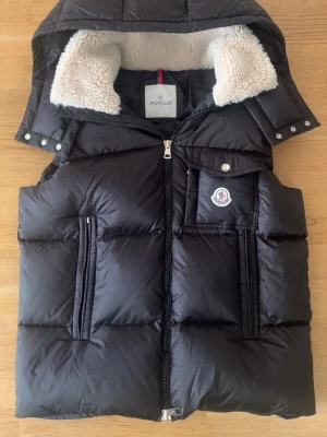 Svart dunväst från Moncler - Snygg svart dunväst från Moncler med avtagbar huva och vit teddyfoder i kragen. Västen har två dragkedjefickor fram och en bröstficka med Moncler-logga. Tillverkad i glansigt material och fylld med dun för extra värme. Perfekt för kalla dagar.