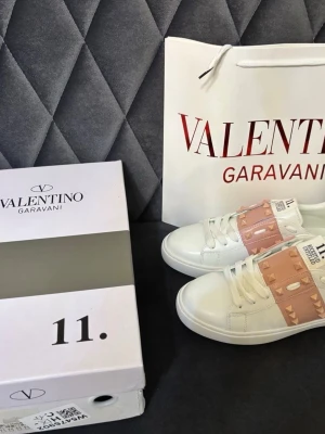 Valentino Garavani vita sneakers - Snygga vita sneakers från Valentino Garavani med rosa band över vristen dekorerat med guldiga nitar. Skorna har klassisk låg profil, vita snören och detaljerade sömmar. Perfekta för dig som vill sticka ut med exklusiv design och coola detaljer.