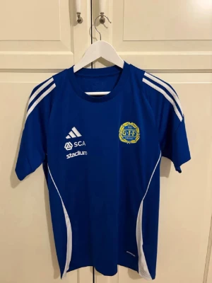 Blå Adidas GIF fotbollströja - Säljer en blå fotbollströja från Adidas med GIF-märke på bröstet och vita detaljer. Tröjan har tryck från SCA, Stadium och McDonald's samt klassiska Adidas-ränder på axlarna. Perfekt för träning eller match. Tillverkad i funktionsmaterial.