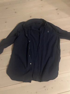 Mörkblå skjorta från Ralph Lauren - Klassisk mörkblå skjorta från Ralph Lauren med broderad logga på bröstet. Skjortan har långa ärmar, knappar framtill och en tidlös design som passar till det mesta. Perfekt för dig som gillar stilrena plagg.