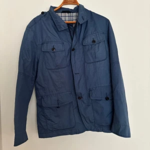 Blå vindjacka med fickor - Snygg blå vindjacka från Lands' End med fyra framfickor och knappar. Jackan har klassisk krage och rutigt innerfoder. Perfekt för dig som gillar en stilren och funktionell look. Längd: 72cm, P2P: 48cm