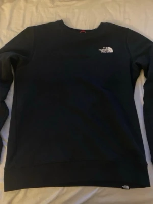 Svart sweatshirt från The North Face - Snygg svart sweatshirt från The North Face med rund hals och logga på bröstet. Tröjan har en klassisk passform och är tillverkad i mjukt material som är skönt mot huden. Perfekt för en avslappnad stil och enkel att matcha med jeans eller joggers.