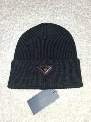 Svart ribbstickad mössa från Prada - Snygg svart ribbstickad mössa från Prada med ikonisk metalltriangel framtill. Klassisk uppvikt kant och diskret logga. Perfekt accessoar för dig som vill ha en stilren och exklusiv look. Materialet känns mjukt och håller värmen bra.