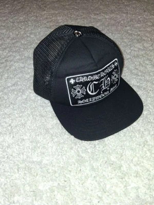 Svart Chrome Hearts truckerkeps - Säljer en svart truckerkeps från Chrome Hearts med vit brodyr framtill där det står 'Chrome Hearts Hollywood USA' och två korsdetaljer. Kepsen har mesh på sidorna och justerbar snapback. Perfekt för dig som gillar streetwear och vill sticka ut.