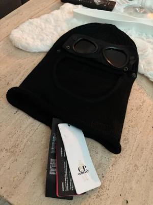 Svart balaclava med goggles  - Säljer en svart balaclava med inbyggda goggles och ribbstickad design. Balaclavan har en öppning för ansiktet och detaljerade sömmar. Perfekt för dig som vill sticka ut med en unik accessoar.