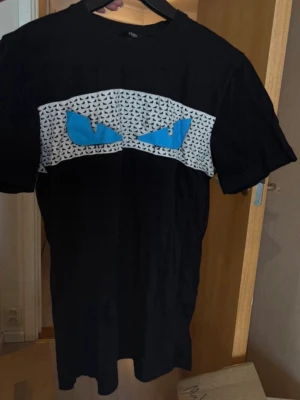 Svart Fendi T-shirt - Cool svart t-shirt med ett unikt tryck framtill som föreställer blå ögon på ett vitt och svartmönstrat band. T-shirten har klassisk passform och korta ärmar. Perfekt för dig som vill sticka ut med en edgy look.