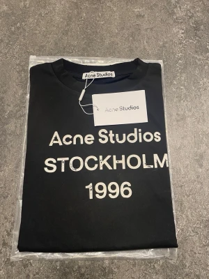 Svart Acne Studios långärmad t-shirt  - Svart långärmad t-shirt från Acne Studios med vit text 'Acne Studios STOCKHOLM 1996' på bröstet. Klassisk rund halsringning och korta ärmar. Perfekt för dig som gillar stilrena och trendiga plagg med tydlig logga.