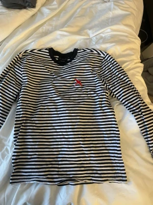 Randig långärmad tröja Axel Arigato - Säljer en svartvit randig långärmad topp med en broderad röd fågel på bröstet. Toppen har rund halsringning och är gjord i mjuk bomull, perfekt för en chill och stilren look. Storlek: M