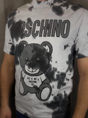 Moschino t-shirt med björntryck - Cool t-shirt från Moschino med stort svartvitt björntryck och logga framtill. T-shirten har korta ärmar och ett unikt batikmönster i svart och grått på vit botten. Perfekt statement-plagg för dig som gillar streetstyle och trendiga prints.