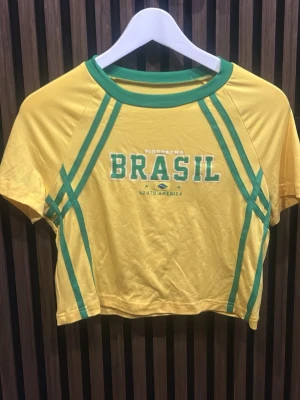 Gul croppad Brasil topp - Sportig gul croppad topp med gröna detaljer och texten 'BRASIL' framtill. Kortärmad modell med grönt kantband vid halsen och gröna linjer längs sidorna. Perfekt för dig som vill ha en färgstark och unik look.
