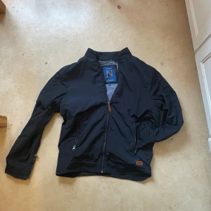 Mörkblå vindjacka nimbus - Snygg mörkblå vindjacka från  med hög krage och dragkedja framtill. Jackan har två snedställda fickor med dragkedja och diskret logopatch vid nederkanten. Tillverkad i lätt och slitstarkt material, perfekt för blåsiga dagar.