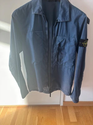 Blå overshirt från Stone Island - Snygg blå overshirt från Stone Island med klassisk patch på ärmen. Jackan har två bröstfickor, varav en med dragkedja, och en rak passform. Alla tillbehör ingår som när jag köpte den. Den är knappt använd. Köptes på Stone Islands hemsida för 2 år sen.