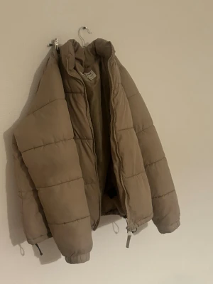 Beige pufferjacka från Denim Day - Säljer en beige pufferjacka från Denim Day i storlek medium. Jackan har hög krage, dragkedja framtill och elastiska muddar vid ärmslut. Perfekt för kalla dagar med sin vadderade och oversized look. Enkel att matcha med olika outfits.