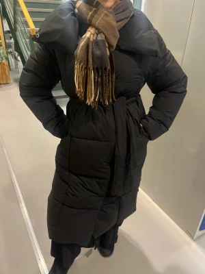 Lång svart pufferjacka med bälte - Säljer en lång svart pufferjacka med bälte i midjan. Jackan har en quiltad design och är riktigt tjock och varm, perfekt för kalla dagar. Stängs med knappar och bälte för extra komfort. Passar dig som vill ha en stilren och mysig vinterjacka.