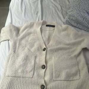 Beige kofta från Vero Moda - Supermjuk beige kofta från Vero Moda med stora knappar framtill och två rejäla fickor. Koftan har v-ringning och är stickad i ett fluffigt material som känns riktigt cozy. Perfekt att slänga på sig över en t-shirt eller linne när det är lite kyligt. Plus så får man med sig en noppborrtagre på köpet!