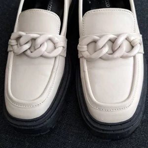 Chunky beige loafers med kedjedetalj - Stilrena loafers i beige skinnimitation från Even&Odd med chunky svart sula och en stor kedjedetalj framtill. Skorna har fyrkantig tå och ger en cool och modern vibe till din outfit. Perfekta för dig som gillar statement-detaljer och trendig design.