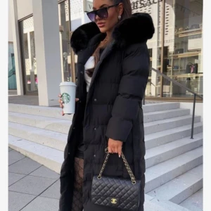 Lång svart dunjacka med fuskpäls - Säljer en lång svart dunjacka med stor krage i fuskpäls och bälte i midjan. Jackan har quiltad design, långa ärmar och stängs med både knappar och bälte. Perfekt för kalla dagar när du vill vara både varm och snygg. Bra skick men har tyvärr ett litet hål går enkelt att sy 💗