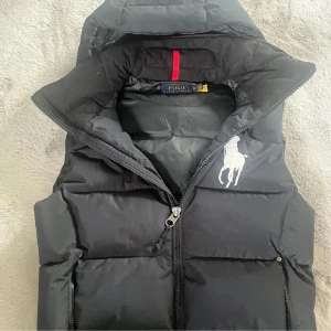 Svart dunväst från Polo Ralph Lauren - Svart dunväst från Polo Ralph Lauren med stor vit broderad logga på bröstet. Västen har hög krage, dragkedja framtill och två fickor med dragkedja. Klassisk puffer-modell som är både snygg och praktisk för kyliga dagar.tveka inte på att fråga på passar till s 