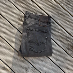 Replay Anbass grå jeans slim fit - Snygga grå Replay Anbass jeans i slim fit med klassisk femficksdesign och svarta detaljer. Jeansen har en diskret tvättad look och är tillverkade i stretchigt jeansmaterial för extra komfort. Perfekta för dig som gillar en modern och stilren vibe.