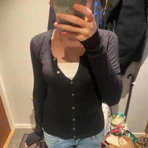 Svart kofta med knappar - En svart, tunn och figurnära kofta med långa ärmar och v-ringning. Koftan har flera små knappar framtill och är enkel i modellen, perfekt att slänga över ett linne eller t-shirt. Materialet känns mjukt och stretchigt.