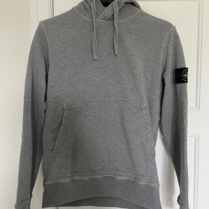 Grå hoodie från Stone Island - Säljer en stilren grå hoodie från Stone Island med klassisk patch på ärmen. Tröjan har huva med snörning, magficka och är tillverkad i mjuk bomull. Perfekt för dig som gillar streetwear och vill ha något bekvämt och snyggt.