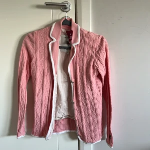 Rosa kofta med vit kant - Snygg rosa kofta från Peak Performance med vit kant längs krage och öppning. Koftan har ett klassiskt kabelstickat mönster och långa ärmar. Perfekt för lager-på-lager och ger en fräsch look till din outfit.