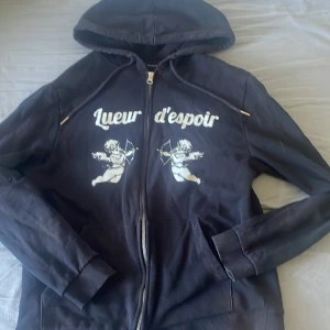 Svart hoodie med tryck från Blinglucid - Svart hoodie från Blinglucid med dragkedja, huva och snörning. Framsidan har ett vitt tryck med texten 'Lueur d'espoir' och två änglar med pilbåge. Mjuk insida och ribbade muddar. Perfekt för dig som gillar streetwear vibes. Storlek M, men passar mindre! Tröjan är exclusiv och säljs inte längre!