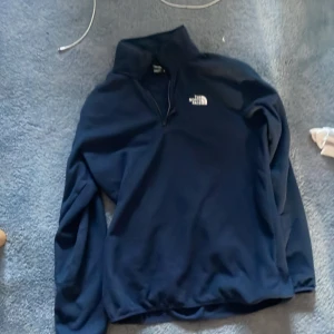 Marinblå fleece-tröja från The North Face - Säljer en marinblå fleece-tröja från The North Face med halv dragkedja och hög krage. Tröjan har långärmade ärmar och en vit logga på bröstet. Perfekt för kyliga dagar och outdoor-häng.