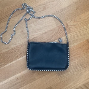 Svart axelväska med kedja och nitar - Cool svart axelväska i skinnimitation med silverfärgad kedjerem och dragkedja upptill. Väskan har rundade hörn och är dekorerad med silvriga nitar längs kanterna för en edgy look. Perfekt storlek för det viktigaste och enkel att bära crossbody. Köpt från zalando