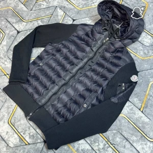 Svart Moncler pufferjacka med huva - Svart pufferjacka från Moncler med stickade ärmar och huva. Jackan har dragkedja framtill, ribbade muddar och Moncler-logga på ena ärmen. Fodret är quiltat och insidan har en cool serietidningsdetalj. Perfekt för dig som vill ha en snygg och sportig look.