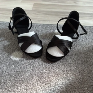 Svarta (latinodans) klackskor med låg klack - Stilrena svarta klackskor med korsade remmar framtill och justerbar ankelrem med spänne. Skorna har en låg, bred klack (5cm) och är tillverkade i ett glansigt satintyg som ger en elegant känsla. Perfekta för dig som vill ha både stil och komfort. Ett litet skav på ena klacken men inget man ser