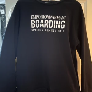 Svart sweatshirt Emporio Armani, - Svart sweatshirt från Emporio Armani med vit text 'BOARDING SPRING/SUMMER 2019' på bröstet. Klassisk rund halsringning och långa ärmar. Perfekt för dig som gillar stilrena och trendiga plagg med logga. Storlek M. Köpt för 3300 i Armanis egna butik i Italien.