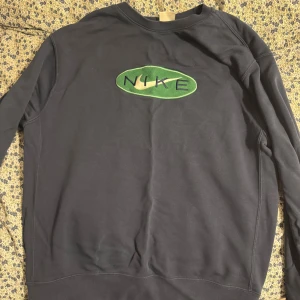 Mörklblå Nike sweatshirt med grön logga. - Cool svart sweatshirt från Nike med en broderad grön och vit logga på bröstet. Tröjan har en klassisk passform och långa ärmar, perfekt för en avslappnad streetstyle-look.