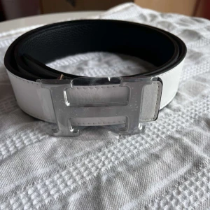 H-bälte - Snyggt bälte i vitt och svart skinn med ikoniskt,H-spänne i silverfärgad metall. Bältet har stilrena, kontrastsömmar och klassisk design som lyfter, vilken outfit som helst. Strl 110cm