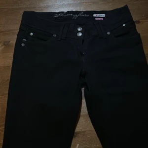 Svarta bootcut jeans från Honeyface - Svarta jeans från Honeyface i bootcut-modell med snygga silverfärgade knappar och klassiska femficksdetaljer. Jeansen har normal passform och är tillverkade i stretchig denim för extra komfort. Perfekta för dig som gillar en tidlös look med lite utsvängda ben.