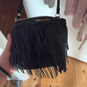 Svart axelväska med fransar - Snygg svart axelväska i skinnimitation med långa fransar framtill och flätat axelband. Väskan har guldfärgad text 'Follow your heart' vid dragkedjan och en kompakt, rundad form. Perfekt för dig som vill sticka ut med coola detaljer.