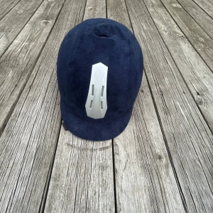 Mörkblå CRW ridhjälm med silverdetalj - Snygg mörkblå ridhjälm från CRW med sammetsliknande yta och cool silverfärgad ventilationsdetalj framtill. Hjälmen har svarta ventilationsöppningar bak och justerbart hakband. Perfekt för dig som vill ha stil på ridbanan.