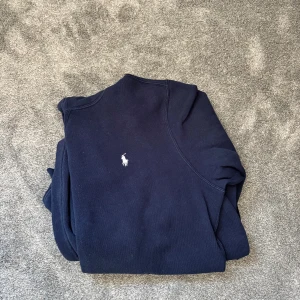 Marinblå hoodie från Polo Ralph Lauren - Snygg marinblå hoodie från Polo Ralph Lauren med klassisk vit broderad logga på bröstet. Tröjan har huva, känguruficka och är tillverkad i mjuk bomullsmix. Perfekt för chill dagar eller när du vill ha en clean och stilren look. Skicket är 10/10, bara använd 3 gånger. 