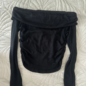 Svart offshoulder långärmad topp - Snygg svart offshoulder topp med långa ärmar och rynkade sidor. Modellen är croppad och sitter tight, perfekt för dig som gillar en stilren och trendig look. Mjuk och stretchig känsla i materialet.