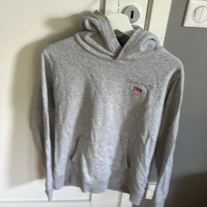 Grå hoodie från GANT med brodyr - Snygg grå hoodie från GANT med broderad logga och sköld på bröstet. Klassisk känguruficka framtill och huva. Mjuk och skön bomullsblandning, perfekt för chill dagar. Passar dig som gillar stilren och sportig look.