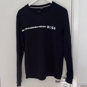 Svart långärmad tröja från BOSS - Mörk blå långärmad tröja från BOSS med stilren silverfärgad logga och linje framtill. Klassisk rund halsringning och normal passform. Perfekt för dig som gillar enkel men snygg design. Materialet känns mjukt och skönt mot huden.