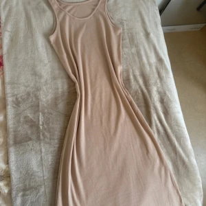 Beige ribbad långklänning - En stilren beige långklänning i ribbat material med ärmlös design och rund halsringning. Klänningen har en figurnära passform och är tillverkad i ett mjukt, stretchigt tyg som känns skönt mot huden. Perfekt för dig som gillar minimalistisk och clean stil.