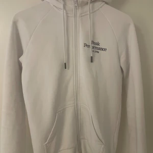 Vit hoodie från Peak Performance - Vit hoodie från Peak Performance med broderad logga på bröstet. Tröjan har huva med snörning, dragkedja framtill och två fickor. Tillverkad i mjuk bomullsmix, perfekt för chill dagar. Klassisk och stilren design med långärmade raglanärmar.
