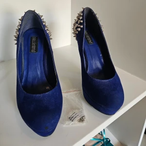 Blå pumps med nitar och strass - Mörkblå pumps från Newtrend i mockaliknande material med hög klack och plattform. Baktill och på klacken är skorna dekorerade med silvriga nitar och strass för en edgy look. Perfekta för dig som vill sticka ut med dina skor.