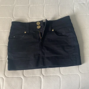 Svart jeanskjol från Gina Tricot - Snygg svart jeanskjol från Gina Tricot med guldiga knappar och dragkedja framtill. Kjolen har klassiska fickor både fram och bak samt en cool detalj med guldig etikett bak. Perfekt för dig som gillar en stilren och enkel look.