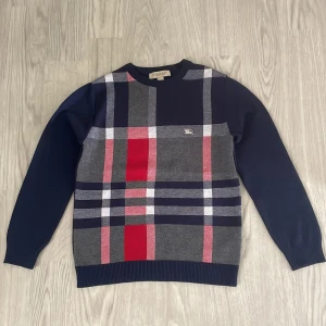 Burberry sweater - Snygg rutig tröja från Burberry London i marinblått, grått, rött och vitt. Klassisk crewneck med lång ärm och broderad logga på bröstet. Tillverkad i mjuk bomull, perfekt för dig som gillar stilren design med en twist. Skulle säga att den är lite liten i storlek och den är i nyskick