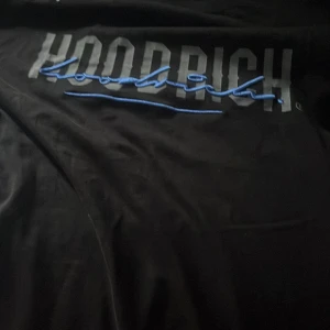 Äkta hoodrich, vet ej va den är värd då ja fick den av en polare - Svart tröja från Hoodrich i streetwear-stil med stor logga framtill i grått och blått broderi. Snygg detalj med patch på ärmen och klassisk crewneck. Perfekt för dig som gillar urban stil och vill sticka ut med cool branding.