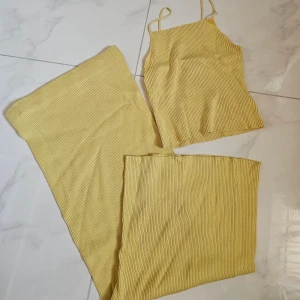 Gul ribbad linnetopp med smala band & kjol - Säljer en ljusgul ribbad linnetopp med smala axelband och rak halsringning. Toppen är ärmlös och har en croppad passform, perfekt för varma dagar. Materialet är mjukt och stretchigt, vilket gör den superbekväm att bära.