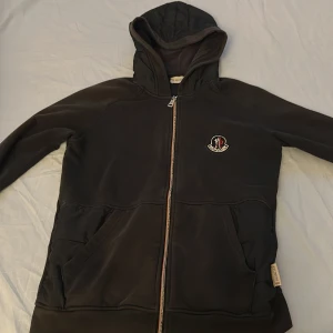Svart hoodie från Moncler med dragkedja - Svart hoodie från Moncler med broderad logga på bröstet och hel dragkedja framtill. Tröjan har två stora fickor, huva och är tillverkad i mjukt material som känns skönt mot huden. Perfekt för dig som gillar stilrena och exklusiva plagg. Helt acceptabelt skick 6,5/10!🔥